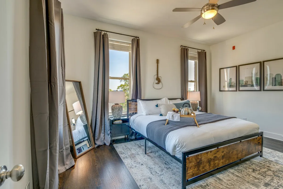 A stylish bedroom with a gray-bedded king bed, a decorative mirror reflecting light, and a guitar on the wall, offering a blend of comfort and unique Nashville flair.