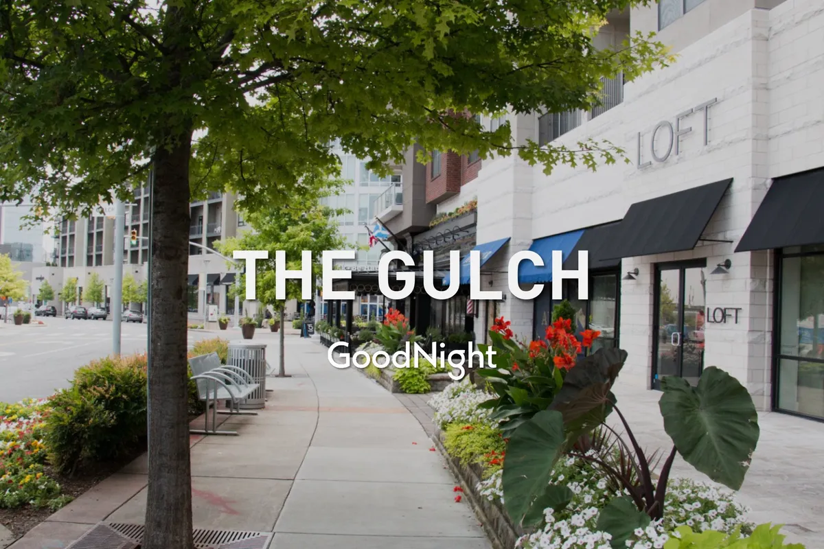 A welcoming streetscape with tree-lined sidewalks, flower beds, storefronts, and benches, capturing the lively and walkable neighborhood vibe. The scene suggests easy access to local shops, dining, and art displays.