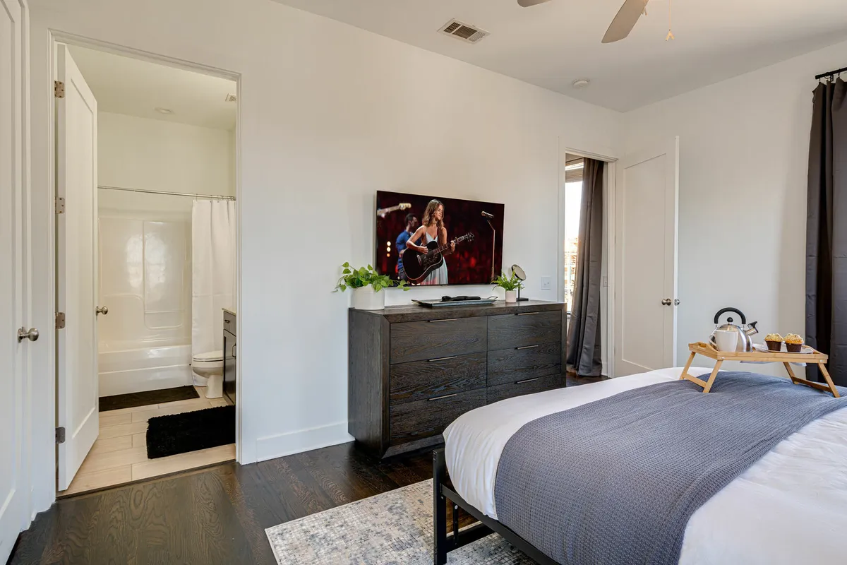 A cozy king bedroom featuring a soft gray bed, a smart TV displaying a musician, a nightstand with snacks and drinks, and vibrant green plants, all set in a comfortable and welcoming atmosphere.