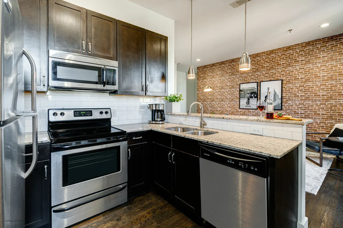 An inviting kitchen space with stainless steel appliances, granite countertops, an exposed brick wall, and pendant lighting. The setup suggests a cozy yet stylish environment suitable for both daily use and entertaining.