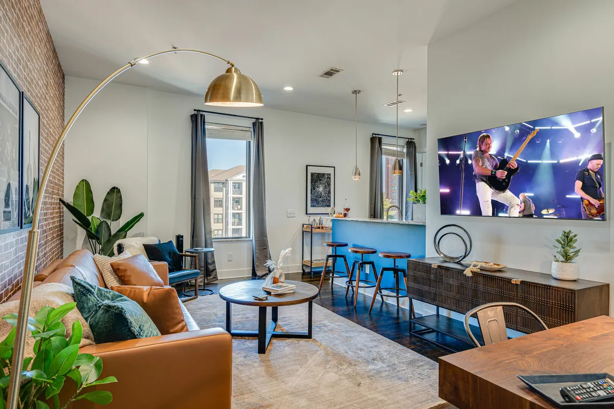 The image captures a chic, open-plan living area featuring a plush leather sofa facing a large TV streaming a concert, a sleek coffee table, vibrant potted plants, and stylish bar stools lined up at a modern kitchen counter. Sunlight streams through the windows, creating an inviting and energetic atmosphere perfect for gathering with friends or relaxing after a day exploring Nashville.