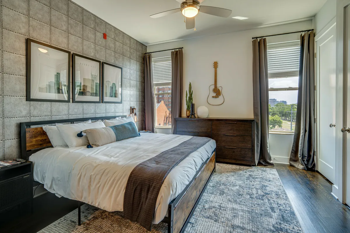 A bright king bedroom with a plush bed layered in soft bedding, eye-catching wall art, a guitar thoughtfully hung on the wall, and sunlight streaming through large windows, creating an inviting and creative retreat.