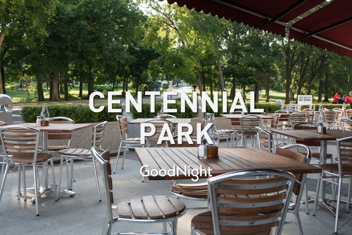 A friendly, relaxed outdoor dining area with wooden tables, metal chairs, and lush greenery in the background. The scene suggests a park-like setting perfect for picnics, casual gatherings, and enjoying community events or sunsets.